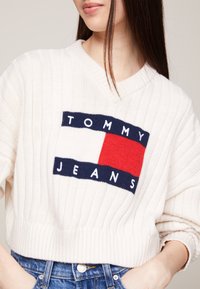 Weißer gerippter Pullover mit V-Ausschnitt und einem blau-weißen-roten Logopatch, auf dem "TOMMY JEANS" über der Brust steht. Kombiniert mit Denim.