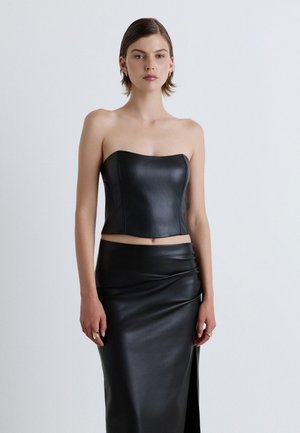 Giovane donna che indossa un top in pelle nero senza spalline e una gonna in pelle abbinata a vita alta con spacco, che sta in piedi davanti a uno sfondo chiaro e uniforme.