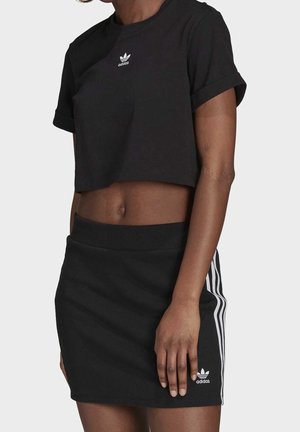 Žena v černém zkráceném tričku Adidas a černé sukni Adidas s bílými pruhy na bocích, záběr od krku po střed stehen.