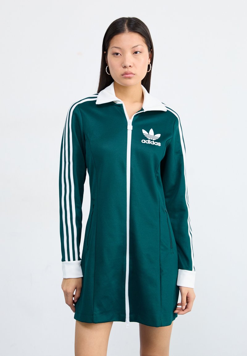 Modell iklädd en grön Adidas zip-up klänning med vita ränder och krage, stående mot en neutral vit bakgrund.