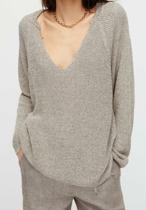 Femme portant un pull beige en maille à col en V avec manches longues et un pantalon en lin taupe, accessoirisée d’un collier chaîne en or.
