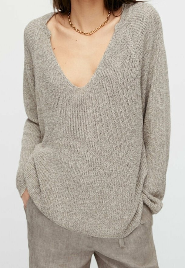 Femme portant un pull beige en maille à col en V avec manches longues et un pantalon en lin taupe, accessoirisée d’un collier chaîne en or.