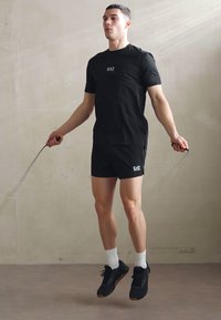 Svart treningst-skjorte og shorts med logo, iført svarte sko og hvite sokker. Personen hopper tau mot en lys vegg.