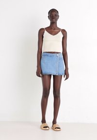 Top alb cu bretele subțiri, fustă mini din denim albastru cu buzunar frontal și sandale bej cu talpă interioară. Textură netedă și design casual.