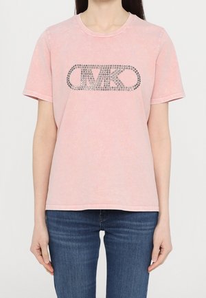 Vrouw draagt een lichtroze t-shirt met korte mouwen en een gespijkerd MK-logo op de borst en een blauwe jeans tegen een effen achtergrond.