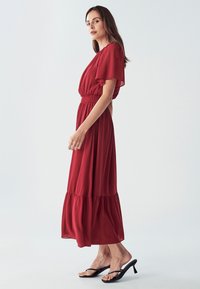 Crvena maxi haljina s kratkim lepršavim rukavima, elastičnom strukom i višeslojnim suknjom. Glatka, lagana tkanina; uparena s crnim sandalama.