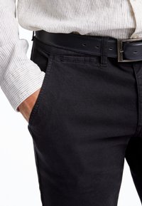 Schwarze strukturierte Hose mit einer Vordertasche, kombiniert mit einem hellen gestreiften Langarmhemd und einem schwarzen Ledergürtel.