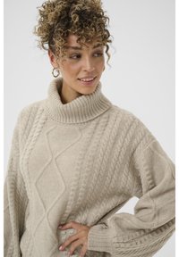 Beige kaapeli-neule turtlenecksweater, jossa on teksturoitu timanttikuosi ja leveät hihat, valmistettu pehmeästä kankaasta mukavan tuntuman saamiseksi.