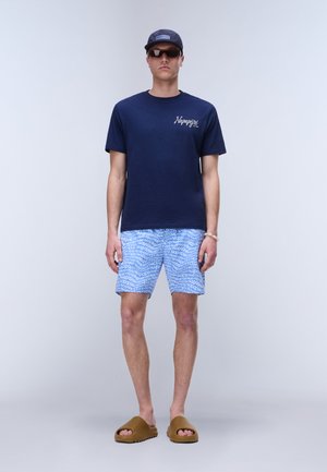 Mand iført navy t-shirt, blå mønstrede shorts med hvid tekst, brune slidesandaler, navy kasket og mørke solbriller, stående mod ensfarvet baggrund.