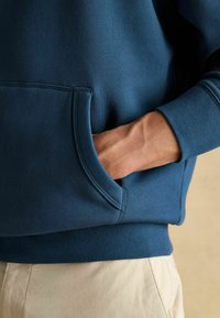 Tume sinine kapuutsiga sviiter, millel on eesmine tasku ja ribakäised. Kangas näib olevat pehme ja sile tekstuuriga, koos heledate beežide pükstega.