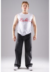 Sleeveless white cotton T-shirt with red scripted text, paired with loose-fitting dark gray denim jeans and white sneakers.