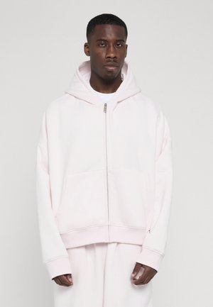 Male model wearing a pale pink zip-up hoodie with a hood, paired with matching pants, standing against a plain light background.