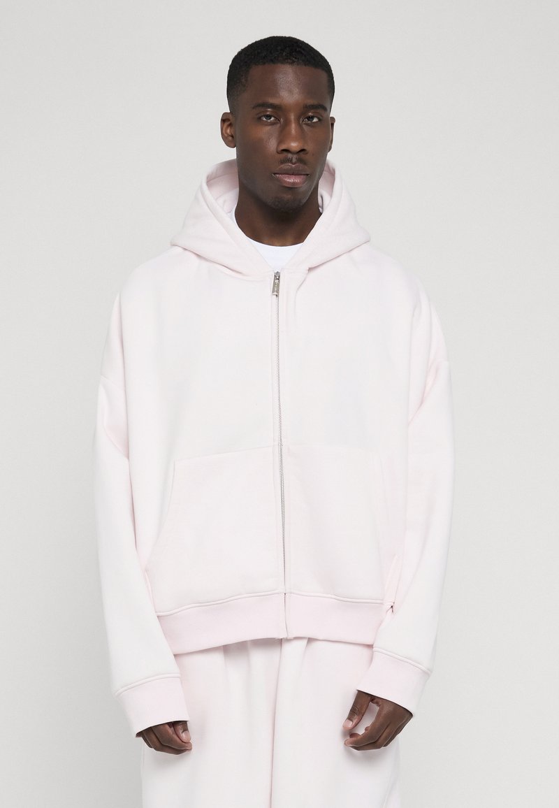 Mannequin masculin portant un sweat à capuche zippé rose pâle avec capuche, assorti à un pantalon assorti, debout devant un fond clair uni.
