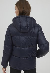Giubbotto puff parmigiano blu navy con cappuccio, caratterizzato da sezioni trapuntate e una texture liscia. Include polsini elasticizzati e un orlo con lieve arricciatura.