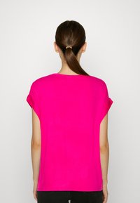 Persona con capelli castani raccolti in una coda di cavallo, che indossa una maglietta rosa brillante a maniche corte e pantaloni neri, di spalle su uno sfondo bianco semplice.