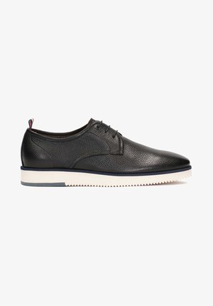 Chaussure noire en cuir texturé à lacets, semelle blanche striée et languette en tissu rouge, blanche et bleue au talon, vue de profil.