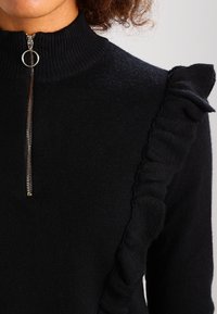 Czarny sweter z wysokim kołnierzem i zamkiem błyskawicznym, z falbaną po jednej stronie. Wykonany z miękkiej dzianiny.