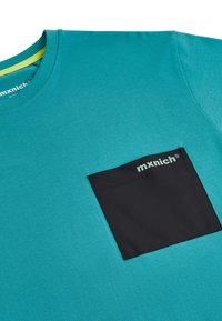 Teal bavlnené tričko s čiernou vreckom a bielym brandingom "mxnich". Okrúhly výstrih, krátke rukávy, jemná textura.