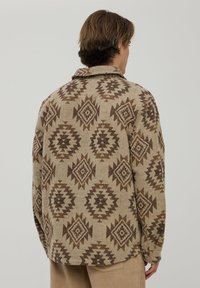 Beżowa kurtka koszulowa z geometrycznym brązowym wzorem w diamenty. Materiał wygląda na strukturalny, z kołnierzykiem i luźnym krojem.