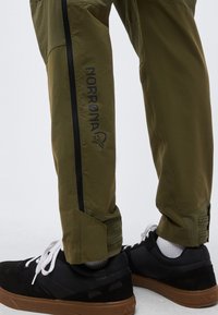 Prim-plan cu pantaloni verde măsliniu cu fermoar negru și cleme cu buclă aproape de glezne, asortați cu adidași negri cu șireturi albe și șosete albe.