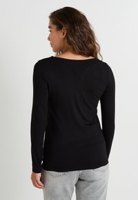 Haut noir à manches longues avec un col rond, fabriqué dans un tissu lisse. Les caractéristiques notables incluent un design ajusté et un ourlet légèrement incurvé.