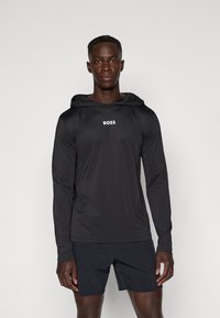 Maglietta a maniche lunghe nera con cappuccio in tessuto leggero, con logo "BOSS" sul davanti. Abbinata a pantaloni corti scuri, con un design aderente.