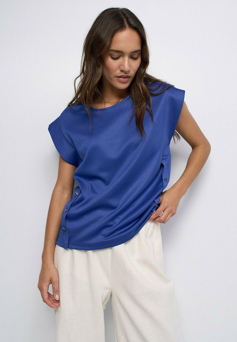 Woman with long brown hair wears a blue sleeveless top with side buttons and loose white pants, standing against a plain light background.
