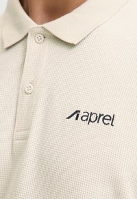 Bežs polo krekls, kas izgatavots no strukturēta auduma, ar apkakli, diviem pogām un tumši zilā krāsā izšūtu logotipu kreisajā pusē.