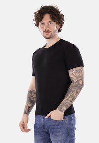 Camiseta negra de manga corta hecha de tela texturizada, con cuello redondo, diseño ajustado, combinada con pantalones vaqueros azules, mostrando tatuajes en los brazos.