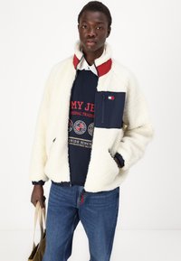 Giacca in pile crema con colletto alto e accenti rossi, dotata di tasca sul petto blu e chiusura con zip. Abbinata a jeans blu.