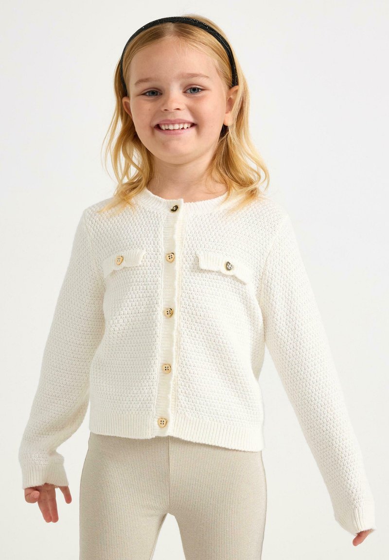 Witte gebreide cardigan met twee voorzakken en houten knopen, gekenmerkt door een textuurpatroon en ribgebreide manchetten. Draagt over beige leggings.