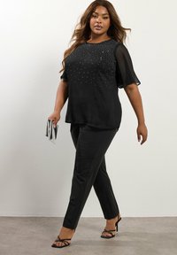 Top nero con maniche corte e trasparenti, decorato con piccoli strass; abbinato a pantaloni neri sartoriali e tacchi a strappo neri.