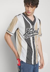 Kortermet V-hals baseballskjorte i stripet beige, grå og hvit stoff, med frontknapper og svart brodert logo.