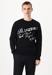 Black knit sweater featuring large white printed signatures and text. Ribbed neckline and cuffs. Smooth texture, classic fit.