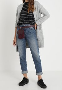 Cardigan de punto gris, camiseta a rayas blancas y negras, jeans de mezclilla azul claro, zapatillas negras y un estuche de accesorio burdeos en un cinturón.