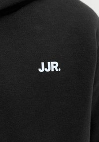 Μαύρο ύφασμα με άσπρα κεφαλαία γράμματα "JJR." τυπωμένα στην περιοχή του στήθους ενός ενδύματος.