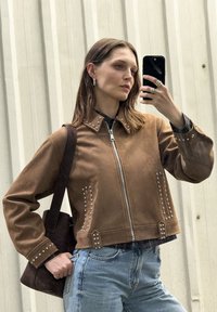 Veste courte en suède marron avec des rivets en argent le long du col et des poches. La mannequin tient un sac tote sombre. Jeans bleu clair et chemise texturée visibles.