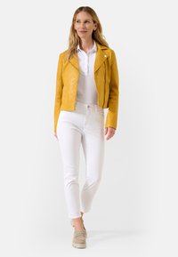 Gele leren motorjack, met ritszakken, licht cropped ontwerp; gecombineerd met witte broek en beige platform schoenen.