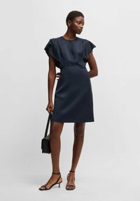 Marinblå klänning med korta utvidgade ärmar, figurnära liv och A-linjeformad kjol. Kombinerad med svarta klackar med remmar och en liten axelväska.