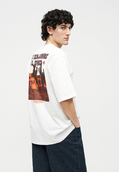 Νεαρός με κυματιστά μαλλιά που φοράει λευκό γραφικό μπλουζάκι με σκηνή δυτικής ερήμου και σκούρο παντελόνι με ρίγες.