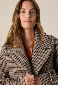 Jeune femme aux cheveux bruns ondulés portant un manteau à carreaux marron sur une chemise orange, tenant le col du manteau avec sa main.