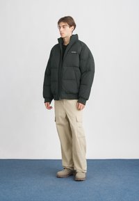 Fiatal férfi fekete pufinak látszó kabátot, bézs cargo nadrágot és bézs csizmát visel, kék szőnyegen áll egy fehér fal előtt.