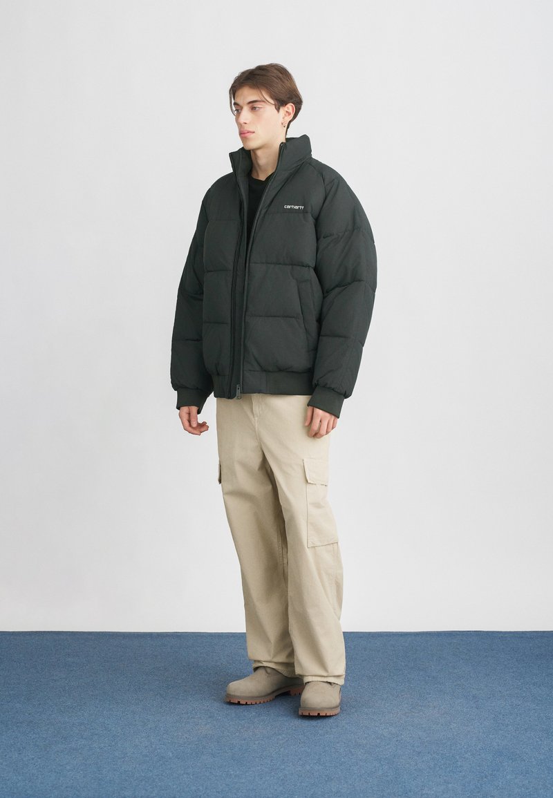 Giovane uomo che indossa un giubbino in piuma nero, pantaloni cargo beige e stivali beige, in piedi su un tappeto blu contro un muro bianco.