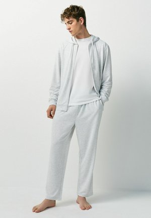 Young man standing barefoot wearing light gray zip-up hoodie, white t-shirt, and matching light gray sweatpants against plain background.