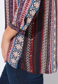 Blusa multicolore con motivi intricati in rosso, blu e crema. Presenta polsini con bottoni e un orlo leggermente curvo.