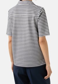 Personne portant une chemise à manches courtes rayée horizontale grise et blanche, debout dos au spectateur et pantalon bleu marine.