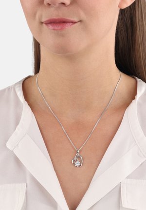 Silberner herzförmiger Anhänger mit kleinen Blumendetails an einer dünnen Kette, die um den Hals einer Frau getragen wird, sichtbar gegen eine weiße Bluse.