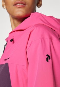 Giacca resistente all'acqua di colore rosa brillante con logo e dettagli neri. Presenta un cappuccio, tasche con zip e una texture elegante e liscia.