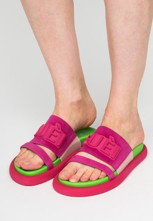 Piedi che indossano sandali aperti davanti di colore rosa acceso e verde con grandi lettere in rilievo sulle fasce, su uno sfondo semplice.