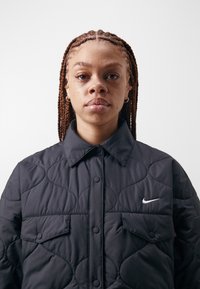 Young person with braided hair wearing a black quilted Nike jacket with buttoned pockets, facing forward against a plain white background.
